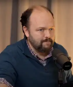 Ross Douthat, wearing a blue sweater and checkered button-down shirt, speaks into a microphone