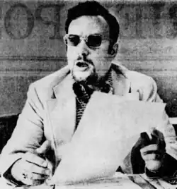 Black and white photo of Frankhouser, wearing sunglasses and holding a piece of paper while speaking