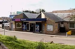 An unusually-shaped yellow-brick building as photographed from slightly higher ground across the road. There is a doorway below a canopy with a sign on it that reads "Royston".