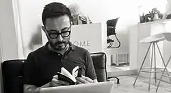 Ruben Quesada sits facing front flipping through pages of an open book. His gaze is down obscuring his eyes.