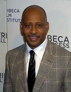 A man in a brown suit with a black tie standing in front of a white backdrop