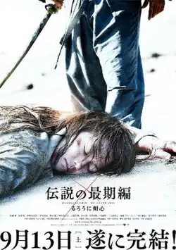 An injured Himura Kenshin lies unconscious on top of sand, while a person wielding a sword stands behind the former's head.