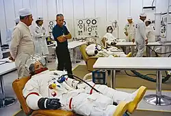 Several men in space suits recline on couches as others stand by