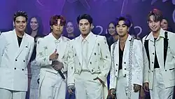 A group portrait of SB19 with each member wearing a white suit wearing a cap and singing to a hand held mic