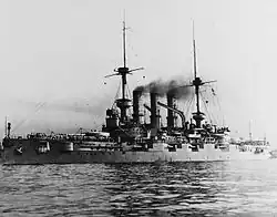 A large gray warship sits in calm water, with dozens of men standing on its main deck.