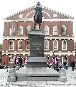 A statue on a pedestal of a man standing with his arms crossed. An inscription on the pedestal reads, "Samuel Adams, 1722–1803. A Patriot. He organized the Revolution and signed the Declaration of Independence." Behind the statue is a three-story brick building with many windows.