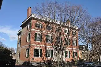 A three story brick house with white trim and black shutters. A wrought iron fence surrounds the front yard, and there is a low railing surrounding the roof.
