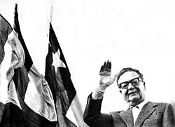 A black and white photograph of Salvador Allende waving. Behind him are Chilean flags.