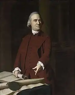 A stern middle-aged man with gray hair is wearing a dark red suit. He is standing behind a table, holding a rolled up document in one hand, and pointing with the other hand to a large document on the table.