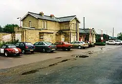 A large two-storey yellow-brick building as photographed from a short distance away. Cars parked in front block the view of the building, and all of the windows are boarded up. In the background lamp posts and overhead gantries on the platforms are visible.