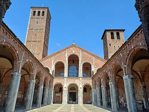 The façade and forecourt of a redbrick church are composed of simple arcades. A brick tower rises up to one side.
