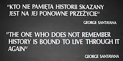 A black placard with white text reading: "KTO NIE PAMIẸTA HISTORII SKAZANY / JEST NA JEJ PONOWNE PRZEŻYCIE" / GEORGE SANTAYANA / "THE ONE WHO DOES NOT REMEMBER / HISTORY IS BOUND TO LIVE THROUGH IT / AGAIN" / GEORGE SANTAYANA