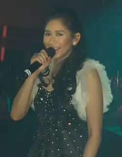 An image of Geronimo in black dress singing while holding a handheld microphone