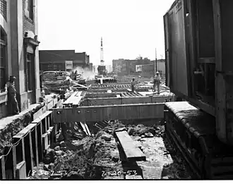 Construction workers observe an open trench formed for a tunnel next to a brick-and-stone building. A concrete and steel framework is being placed over the open trench.