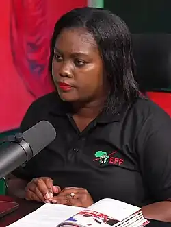 A woman seated at a table and behind a microphone, wearing a black polo shirt