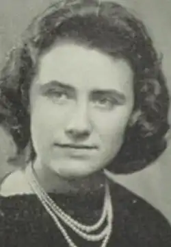 A young woman with light skin and dark curly hair, wearing a dark top and strands of pearls or beads