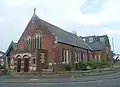 Three-quarter view of a building in red brick and with doubled entrance doors and brick-dressed three-light lancet windows with stone mullions.