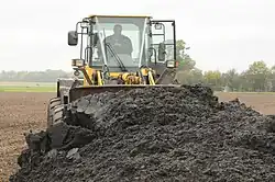 A yellow shovel excavator with a man visible at the wheel in the cab, is pushing a huge pile of sewage sludge which looks like very dark colored dirt, which is filling most of the bottom half of the image. This is taking place on a bare field, with light brown soil visible, whose color contrasts strongly with the dark sewage sludge. In the background, there is a green field, and a yellow field, and trees.