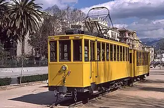 Yellow painted tram and trailer on short section of track. Palm trees behind.