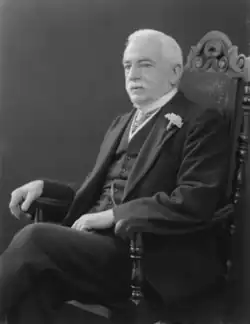 A vintage photograph of an elderly man with white hair and a moustache, wearing a dark suit with a white shirt, a bow tie, and a flower on his lapel, standing and looking slightly to the side.