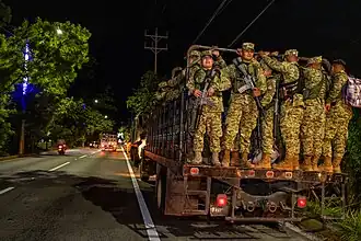 Soldiers in camouflage uniform, armed with rifles, and standing in an uncovered truck in San Marcos
