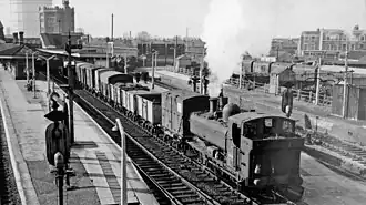 A pannier tank locomotive, travelling in reverse, is pulling a train of open and closed goods wagons through a station.