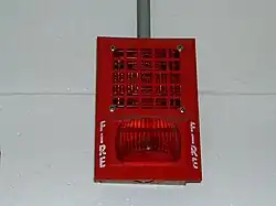 a red device built into a wall. The top half is a speaker and the bottom half is a red light