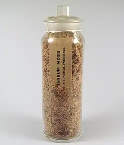 A stoppered glass jar filled with a brown crumbly substance. A card partially visible in the jar reads "...HAGNUM MOSS / ... in surgical dressings."