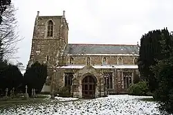 A stone church, with a porch and a square tower with pinnacles, set in a churchyard with trees and a dusting of snow