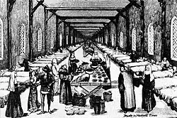 A long hall lined with beds. In the centre is a table with food, surrounded by monks and nuns.