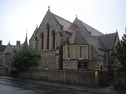 A low-roofed, wide, cobbled flint church with several sections, facing a road behind a wall of the same material. On the right is an apse with plain stone-dressed lancets; next to it is the body of the church, with tall round-headed windows. Beyond that is a spirelet and a low extension with a five-light window.
