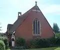 A red-brick church with a tiled roof extending nearly to ground level. Low shrubs surround the building on all sides. A three-light lancet window with stone mullions dominates the nearest side. A small stone cross and bell-tower are on the roof. The left-hand side has a white entrance porch.