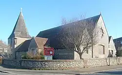 Three-quarter view of a long flint church with an extremely wide, stumpy tower on the left. This is topped with a dumpy grey spire. The brown church roof is heavily discoloured. A tree in full leaf obscures the near corner.