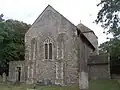 A flint rubble church with large stone quoins. In the nearest wall is a two-light lancet window between buttresses. A squat tower with a squared-off pyramidal roof is partly obscured behind this.