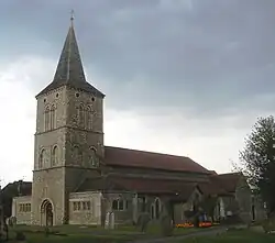 Three-quarter view of a flint church with a tall, three-stage tower to the left, topped with a dark grey spire. Low projections with paired arched windows flank the tower on both sides. A porch juts forward from the main body of the church, next to three low lancet windows. The roofs are of red tiles. Irregularly spaced gravestones are in front of the church.