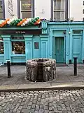 A stone well head mounted in the middle of a footpath in front of a row of businesses on a street. There is plaque about the plaque visible on the wall behind the well.