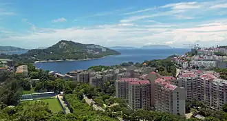An inlet on a seacoast with some islands on the horizon, seen from a height. Directly behind it is a high ridge on a narrow, small peninsula that is mostly wooded with some development visible. In the foreground are medium-height buildings with red roofs on the center and right, a construction crane on the far right, and a playing field on the left.