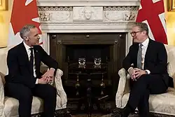 Mark Carney sits and talks with Prime Minister of the United Kingdom Keir Starmer flanked by Canadian and UK flags for each respective country's leader. Both are dressed in a dark suit, white shirt, and tie