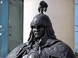 Torso and head of a statue of a man in full ceremonial armour with plume on helmet, in front of a pillar.