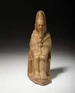 Small yellowish statue of a man seated on a throne, wearing a tight fitting robe, a crook and a flail