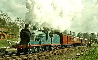 A blue steam locomotive pulling a train of one mail coach and four or five passenger coaches, all painted brown, through Lisburn station.
