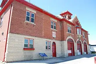 A two-storey red brick and concrete fire hall with three vehicle doors, one regular door, and two windows.