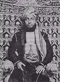 A black-and-white photograph of a man with a dark beard wearing a turban, a dark jacket, and a white shirt, sitting, and looking at the viewer