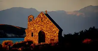 A small sunlit stone church by a lake with mountains in the distance