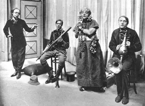 Stage group of four middle-aged white people in extravagant clothing; two of the men hold ornate and improbable musical instruments and the one woman is declaiming