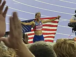 Photo of Sydney McLaughlin-Levrone wearing a crown and holding up an American flag behind her back with spectators in the foreground and with violet athletics tracks in the background