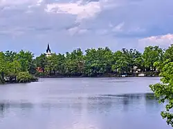 A landscape image; in the foreground can be seen the waters of the Svět pond, with light ripples. Behind them can be seen thick and vibrantly green foliage, out of which rises a steeple from the town of Třeboň. The sky is overcast.