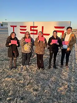 Five workers are standing together while smiling. They are holding signs that say "union" wrapped in hearts. Behind them, is a concrete stand emblazoned with the Tesla logo.