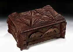 An image of a wooden box taken in 3 quarters view with many textured three dimensional sections created by layered pieces of wood. The top has a star motif, the front two wooden pistols, and the side the state outline of Texas.