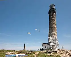 This old photograph shows a stone lighthouse built atop a rocky ledge. It is connected by a covered and enclosed walkway to a house on the left. A small white shed is visible in the foreground.